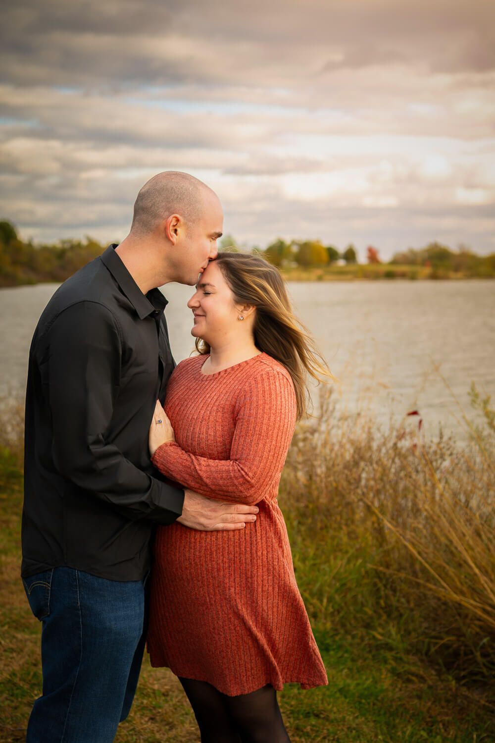A romantic Omaha couple takes photo embracing by a lake at sunset with dramatic cloudy skies and autumn foliage in the background.