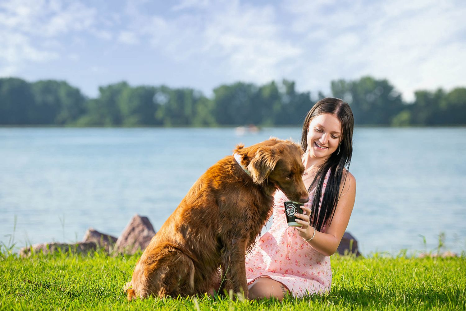 A golden retriever dog and person relax together on green grass near a peaceful lake on a sunny summer day.