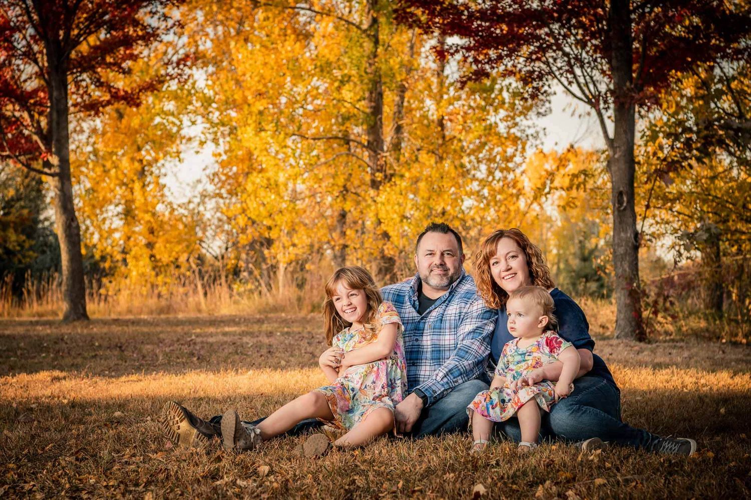 A family sits together on fallen autumn leaves in a park surrounded by golden trees during sunset.