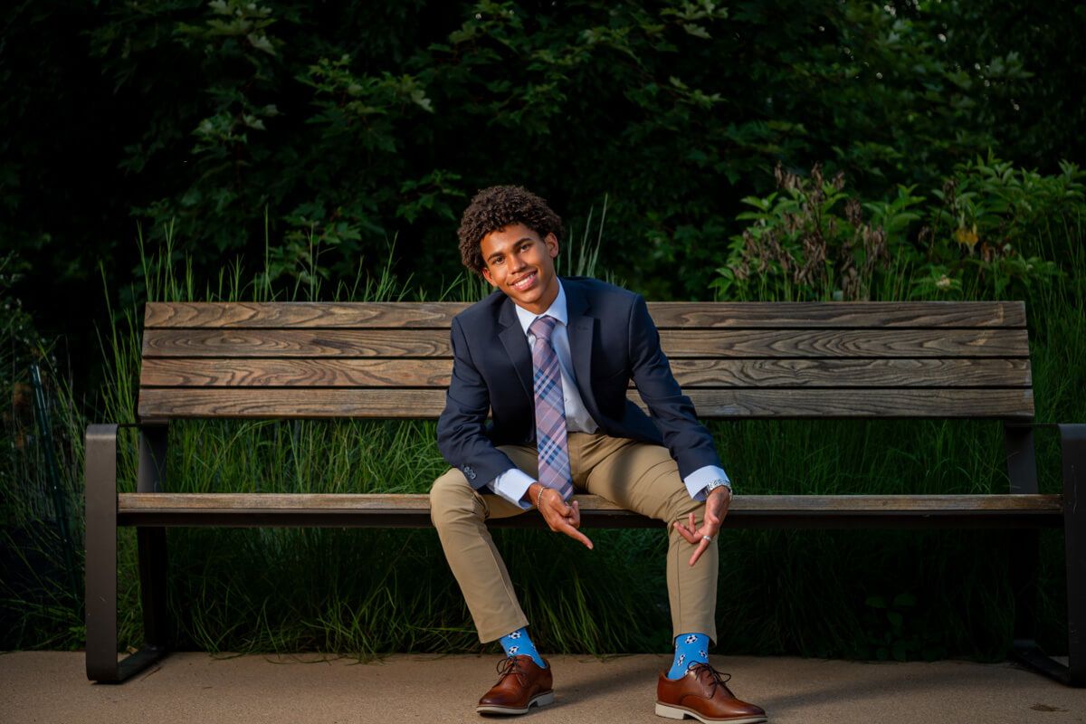 Young man in navy blazer and colorful socks sits confidently on a wooden bench surrounded by lush greenery.