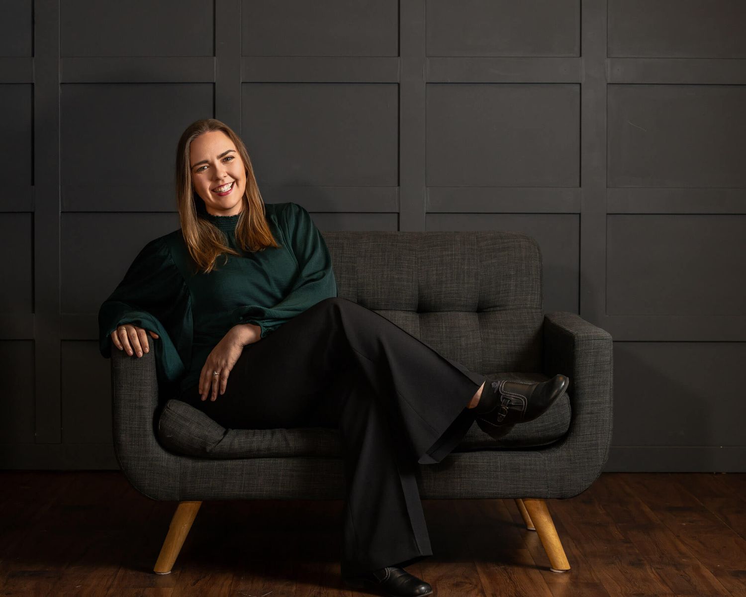 Woman in green top and black trousers smiling on a grey tufted sofa against a dark panelled wall.