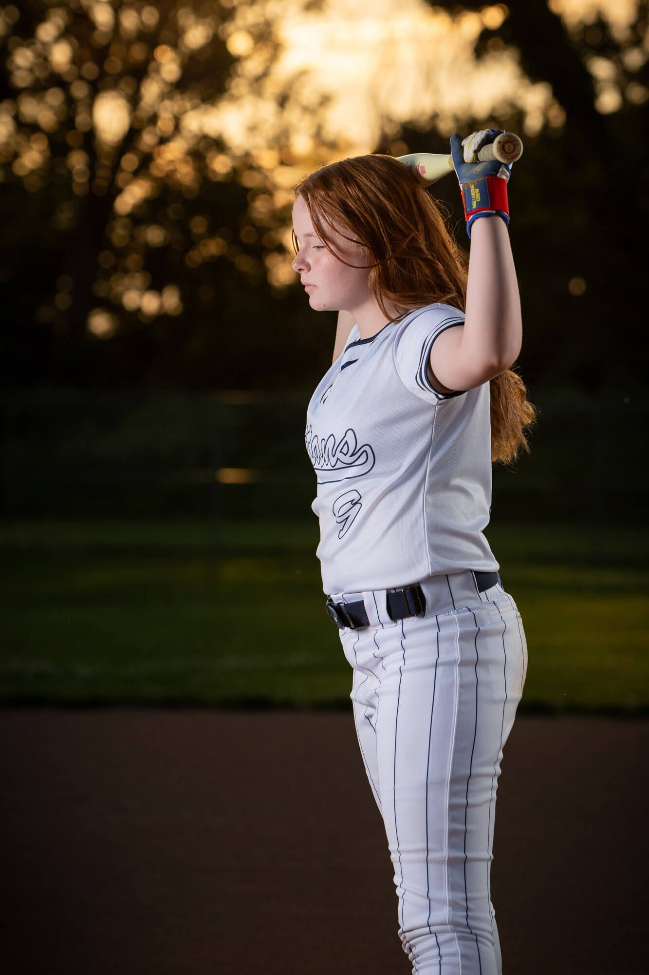 A young female softball player in white uniform holds a bat over her shoulder at golden hour on a field.