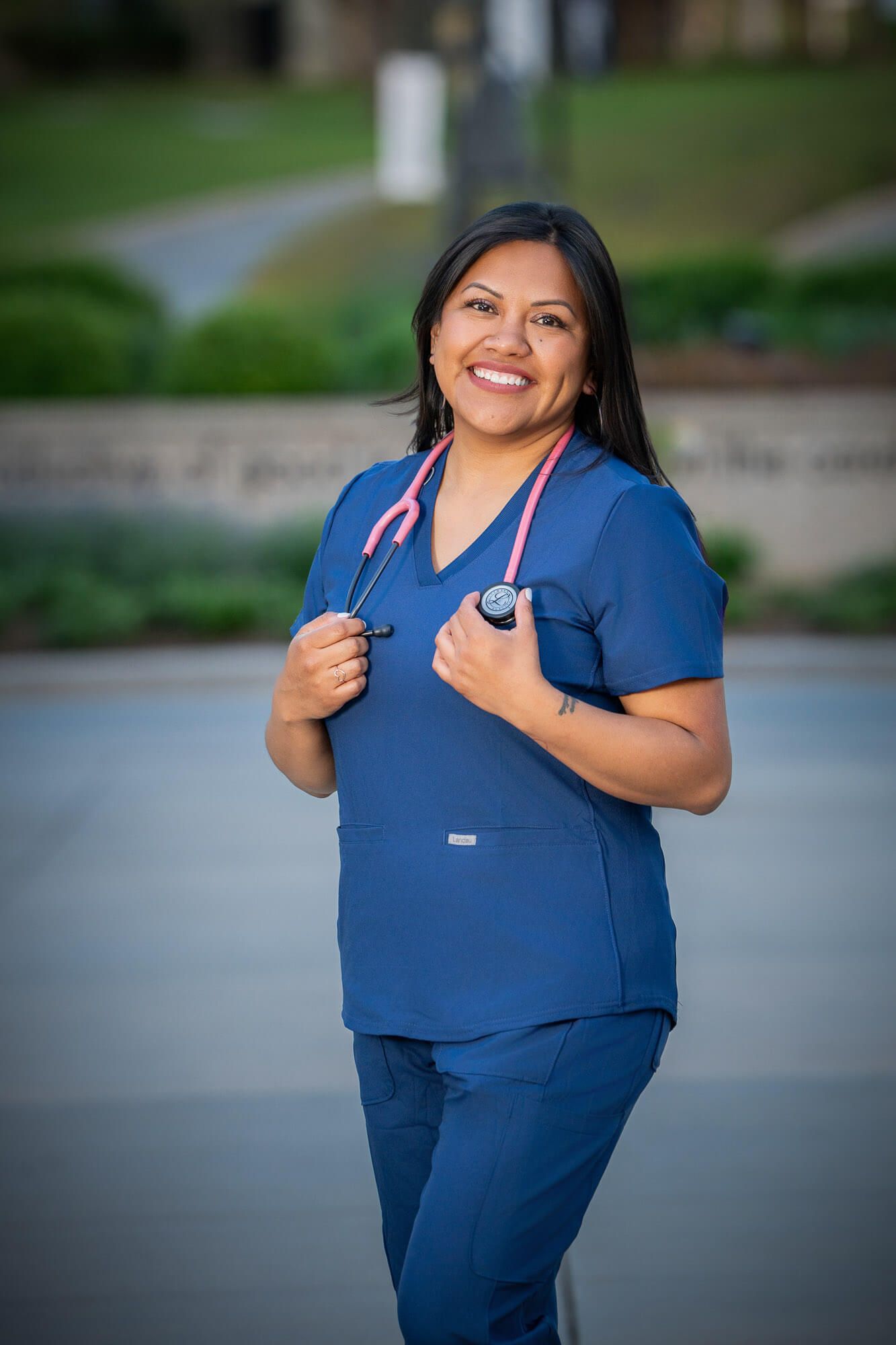 Smiling female nurse in navy blue scrubs with pink stethoscope poses outdoors at medical facility.