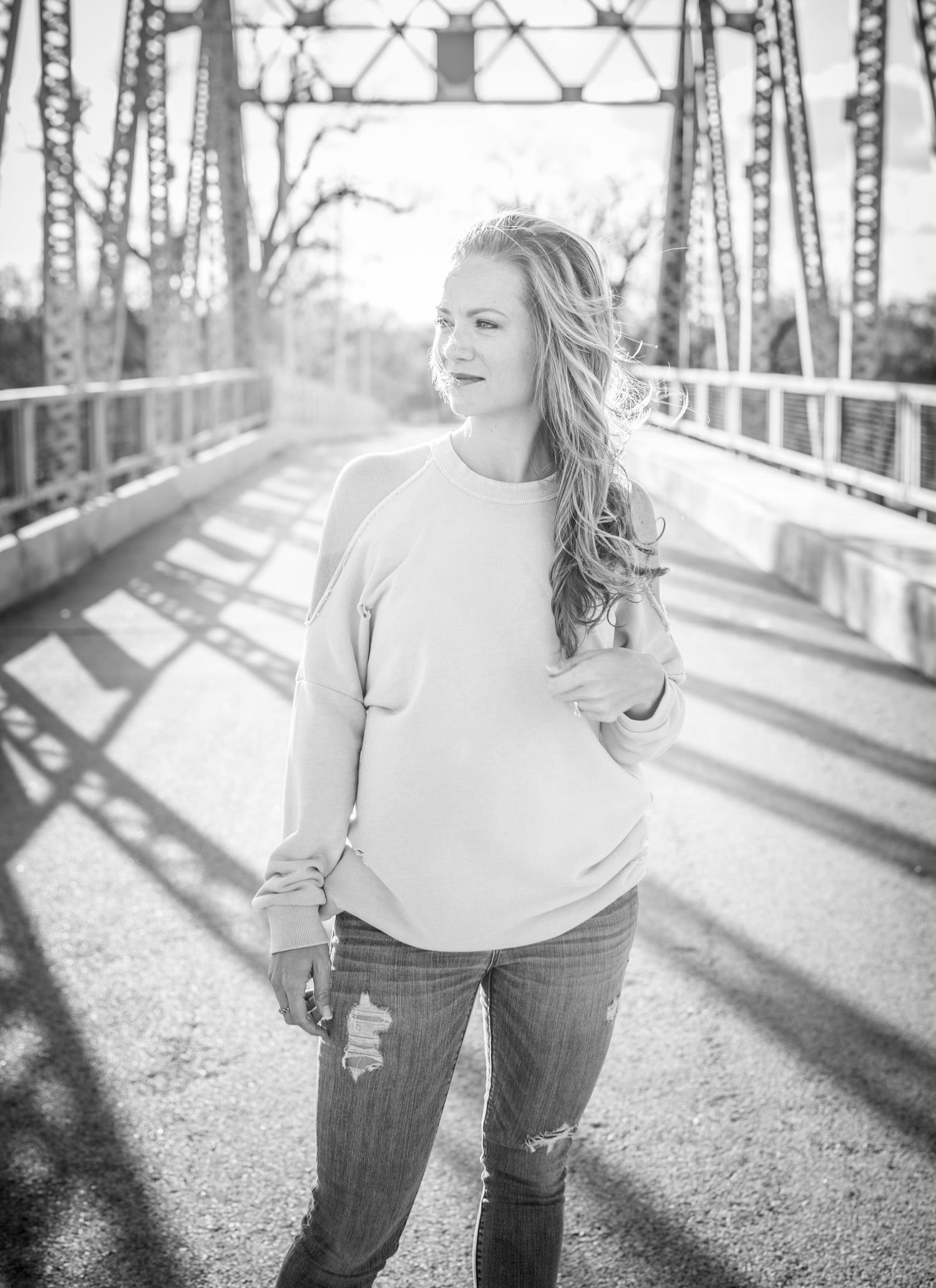 Black and white portrait photograph of a young woman in a sweater and jeans standing on a rustic bridge.