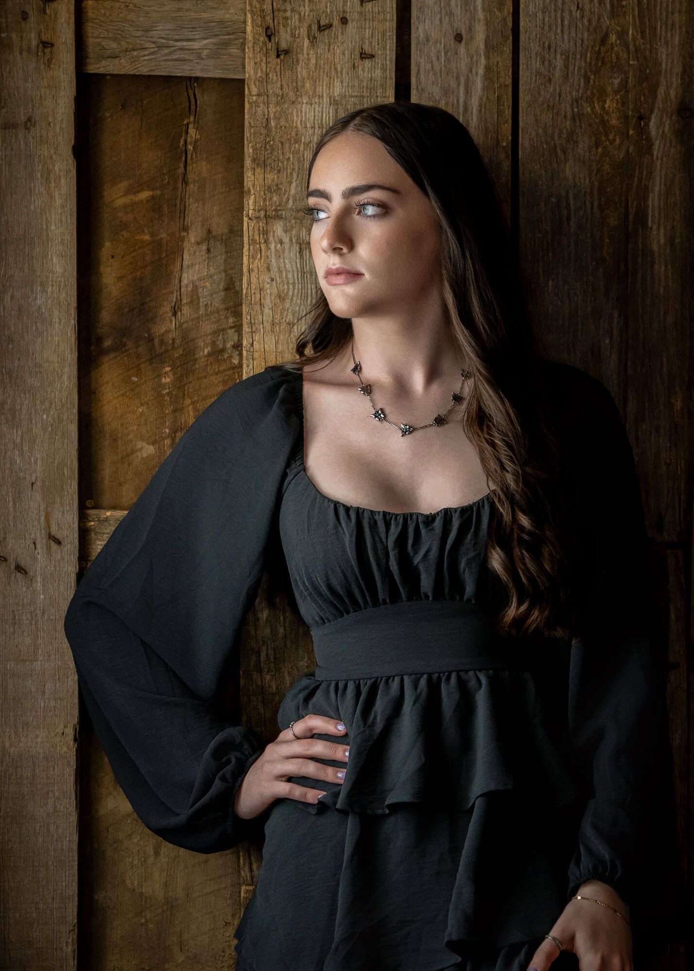 A woman in a black renaissance-style dress poses against a rustic wooden wall in dramatic lighting.