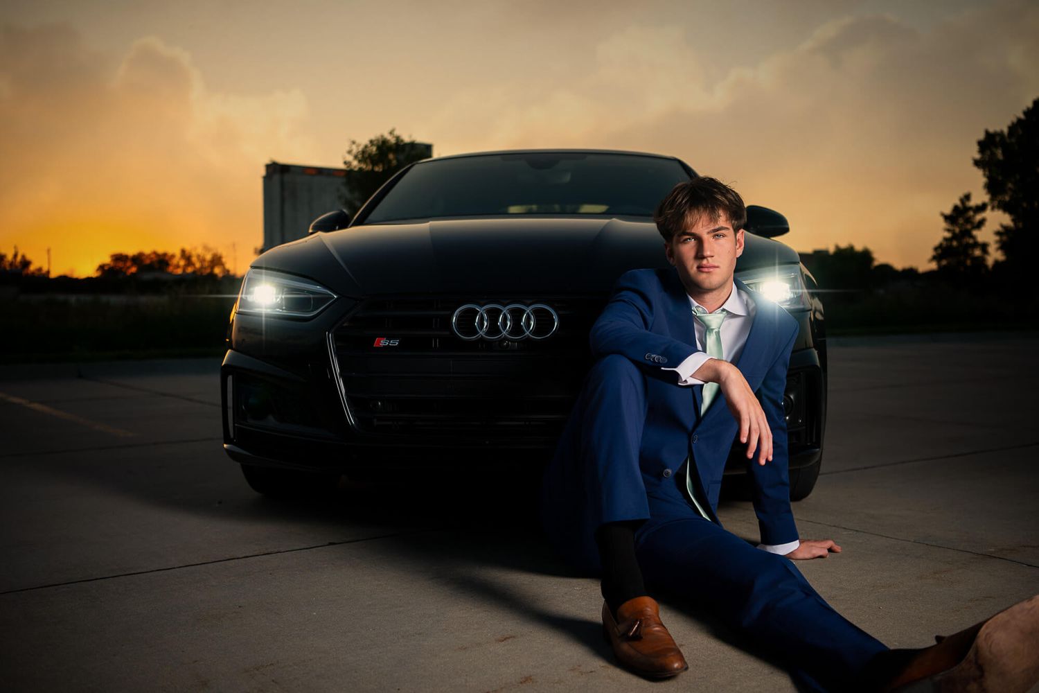 Westside High School senior photo session of a male senior in a blue suit sitting in front of his Audi car at sunset