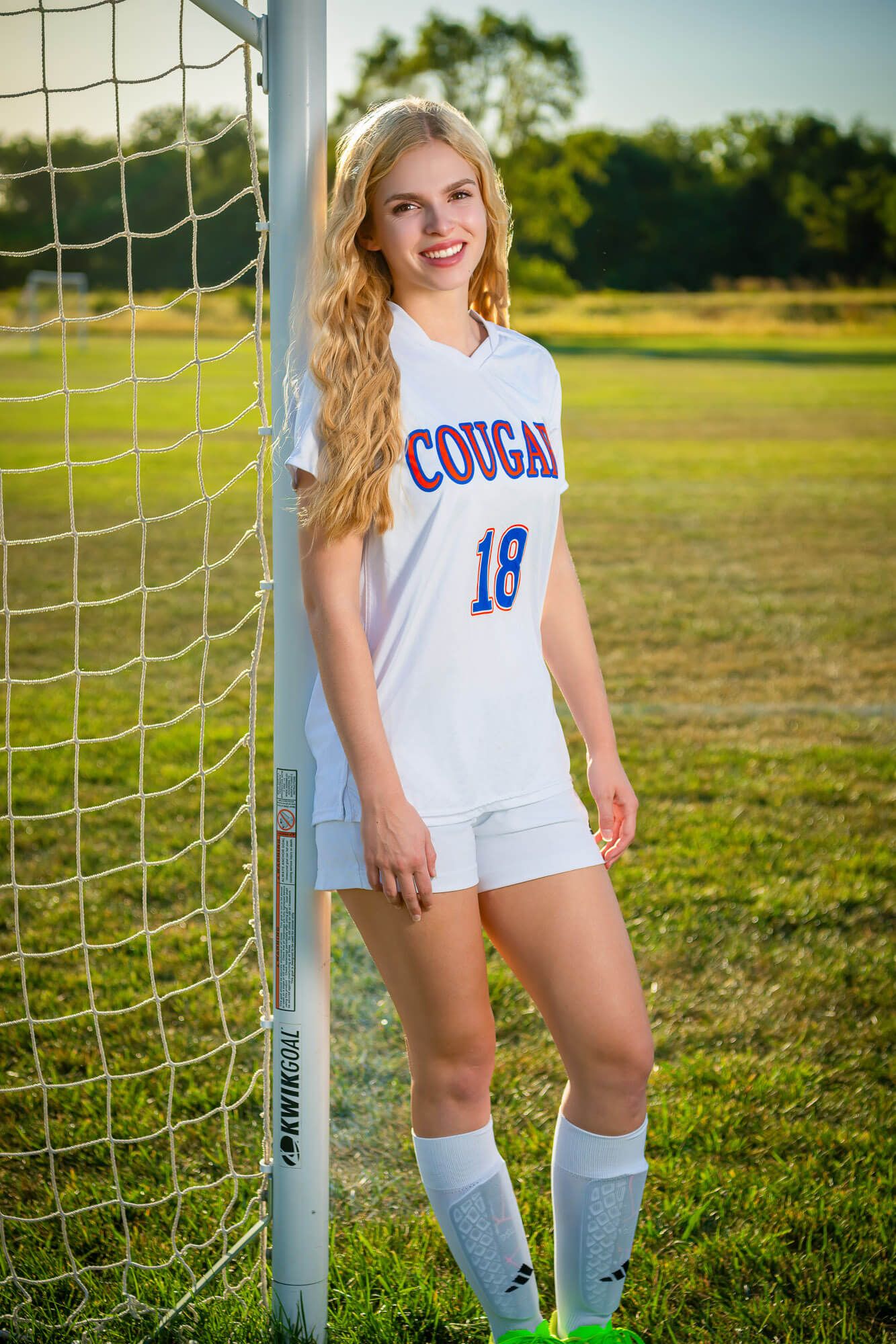 Soccer player poses by goal post wearing white uniform with number 18 on sunny field.