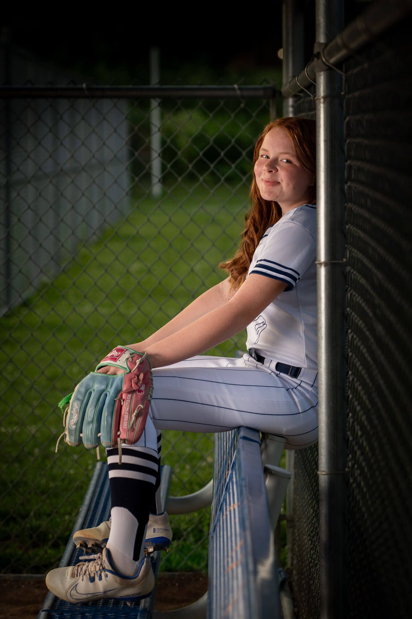 Young redhead girl in white softball uniform sits in dugout holding colorful batting gloves, leaning against fence.