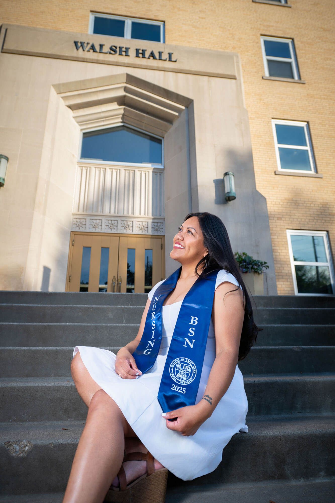 BSN graduate in white dress and blue sash sits on steps outside Walsh Hall, smiling upward in celebration.