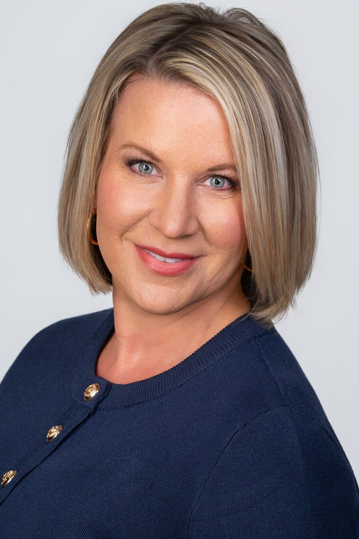 Professional headshot of a smiling blonde woman with blue eyes wearing a navy cardigan with gold buttons.