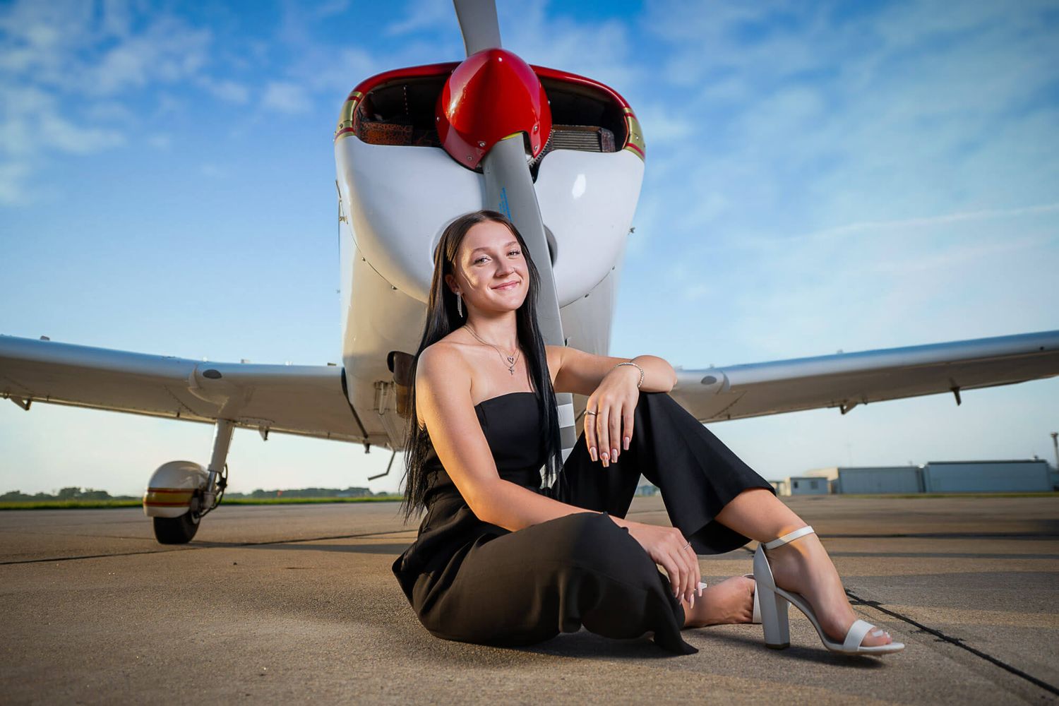 Young woman in black outfit poses gracefully in front of a small red and white propeller airplane on a tarmac.