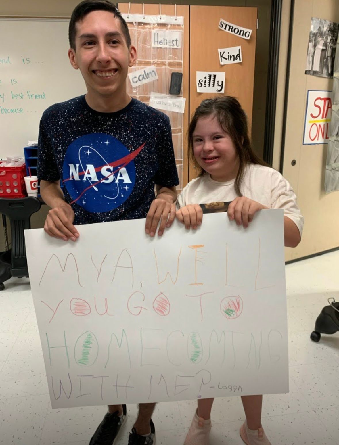 Two people pose together holding a white sign with writing on it in front of a classroom bulletin board.