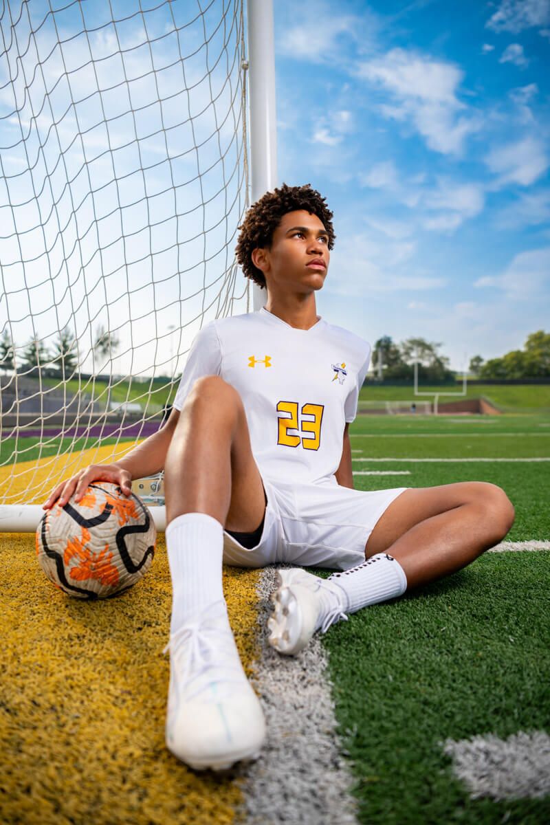 Senior soccer player from Bellevue West High School resting on soccer field near goal post with ball, white uniform, and white cleats.