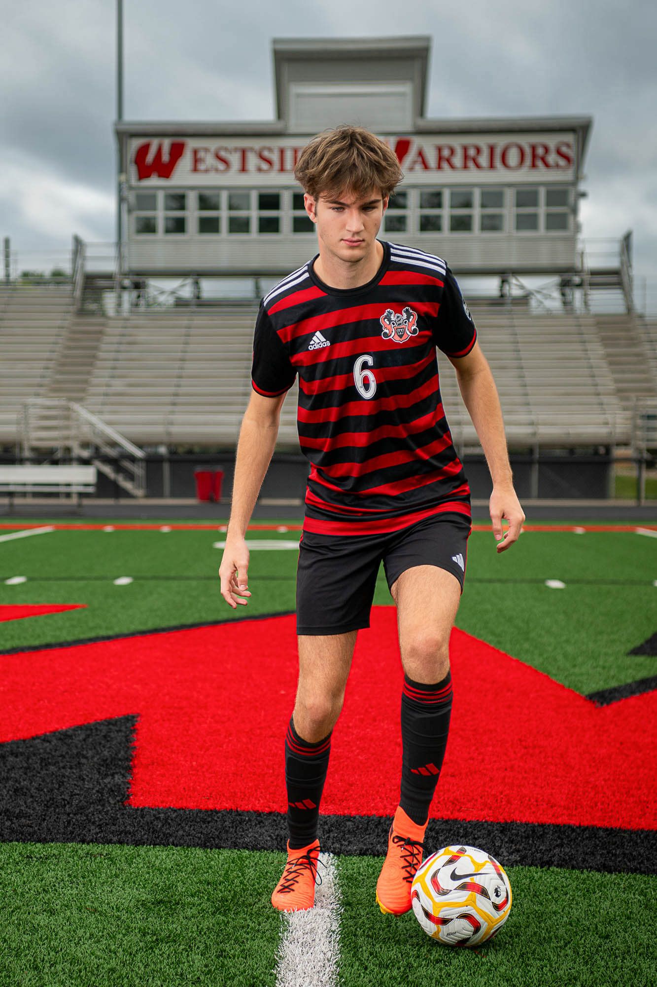 Soccer player in red and black striped jersey poses with ball on field beneath Westside Warriors stadium sign.