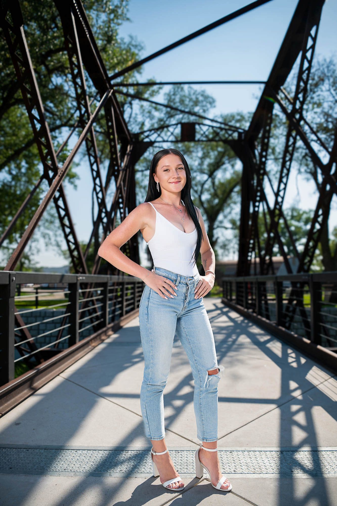 Woman in white tank top and ripped jeans poses confidently on a metal bridge walkway in sunlight.