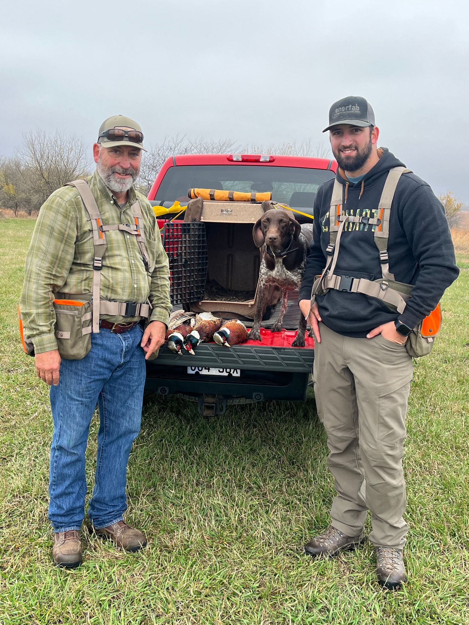 Two hunters stand with their hunting dog and harvested pheasants in the back of a pickup truck on a cloudy day.