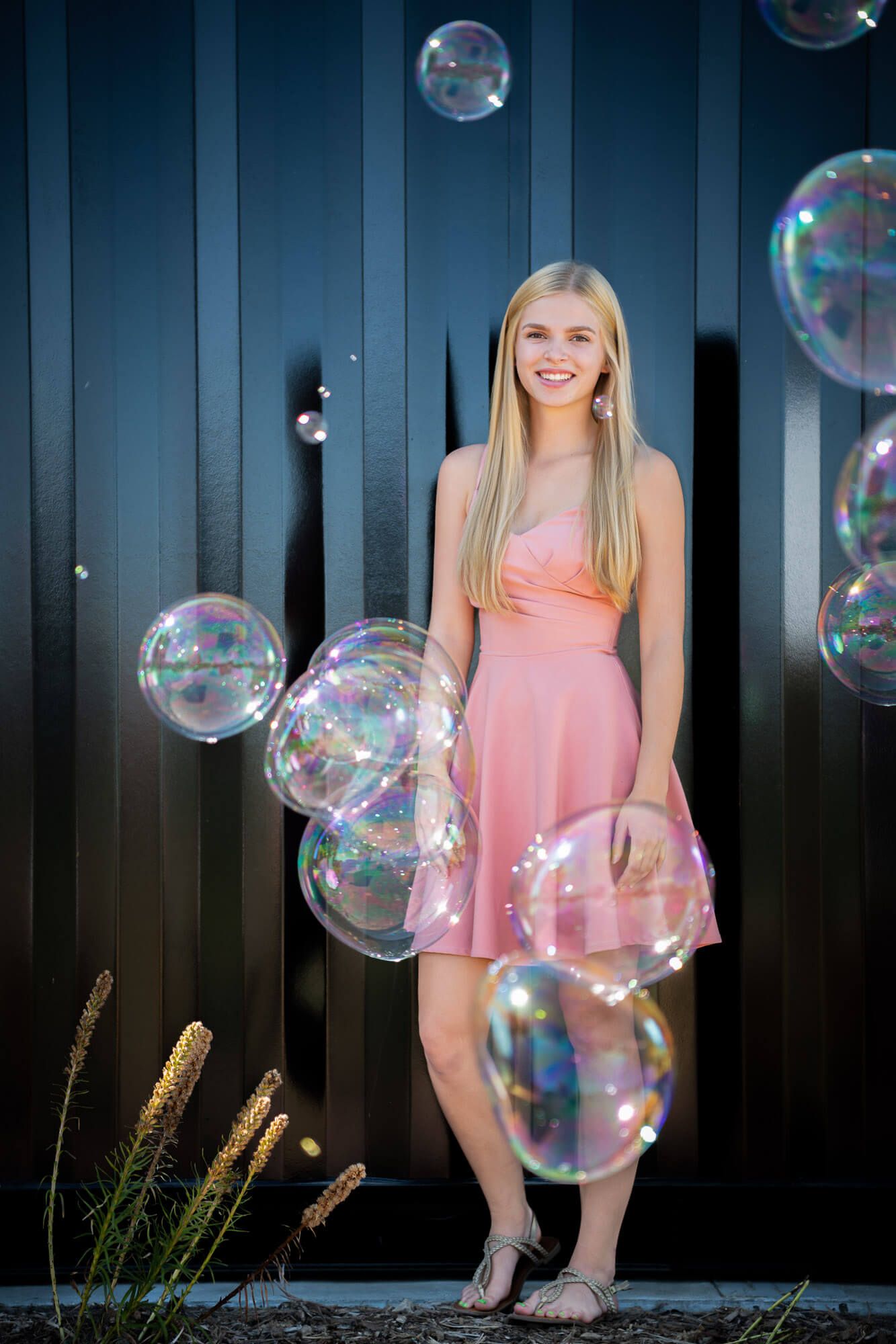Smiling blonde teen in pink dress surrounded by large soap bubbles against dark metal wall.