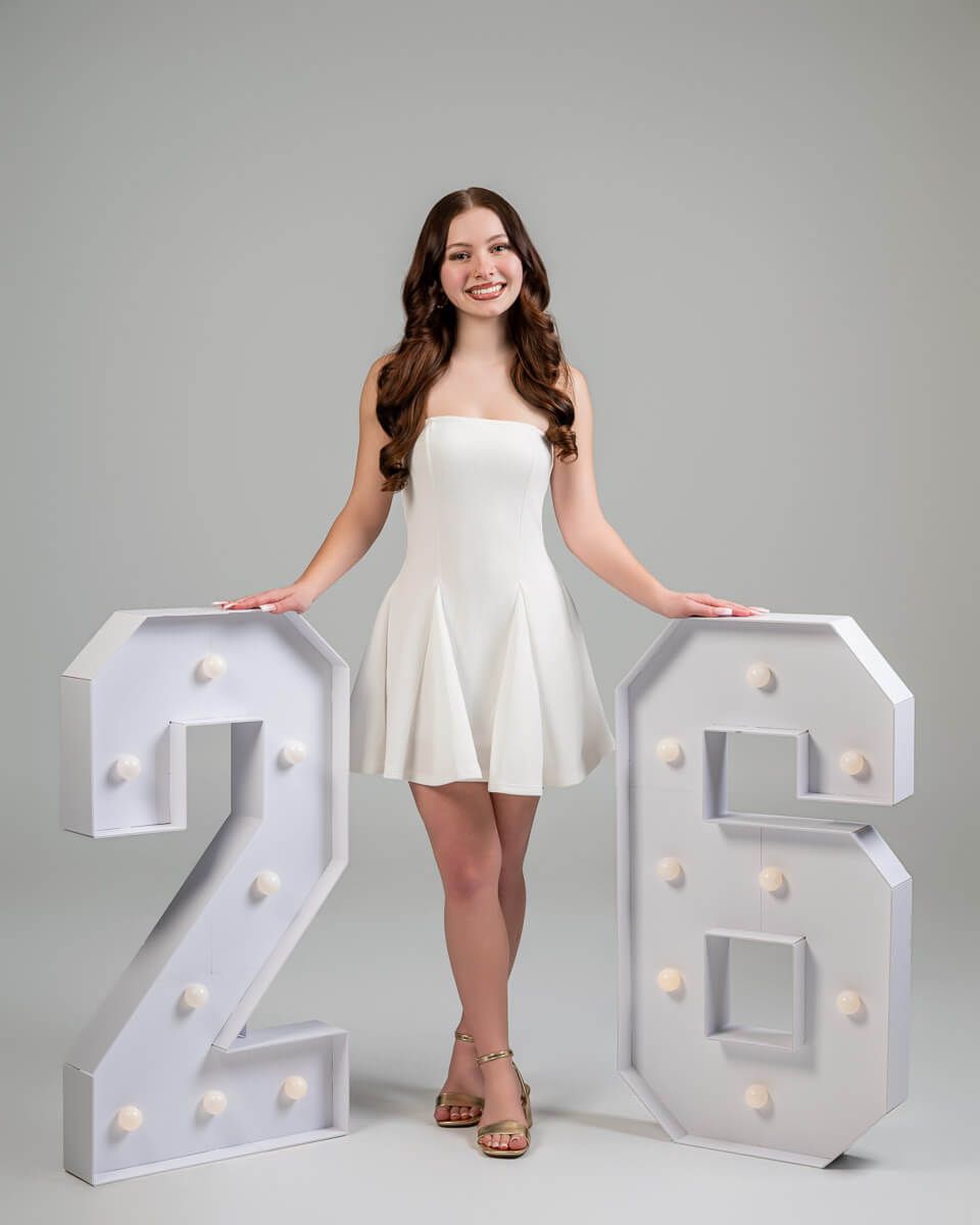 Young woman in white strapless dress poses between large light-up marquee numbers 26 for graduation photos.