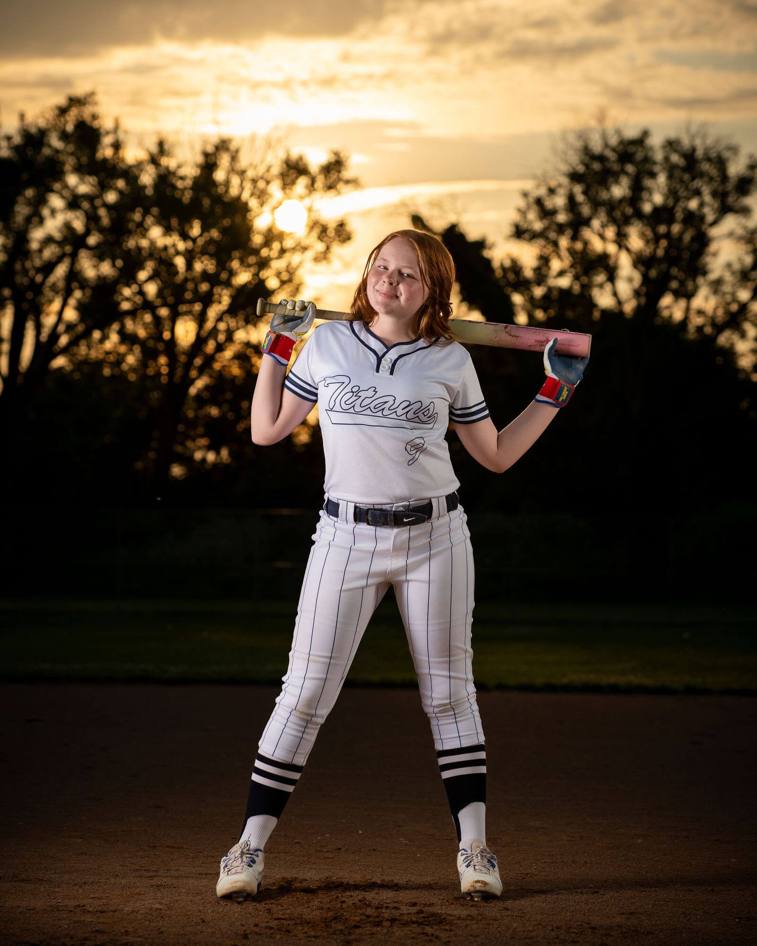 A softball player in a white Titans uniform poses with a bat over her shoulders at sunset on a dirt field.