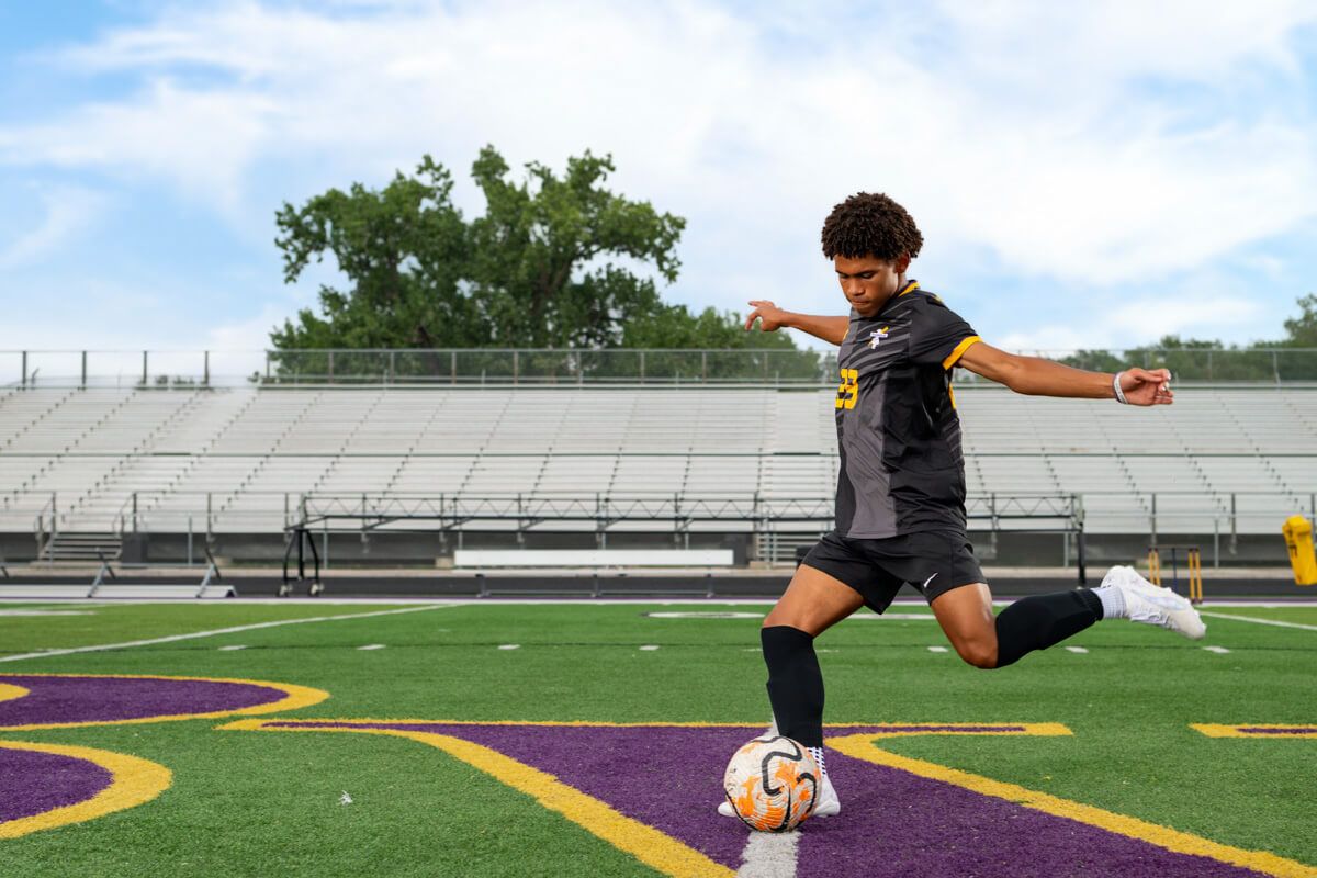 Soccer player in black jersey kicking a ball on a purple and gold turf field with stadium bleachers behind.