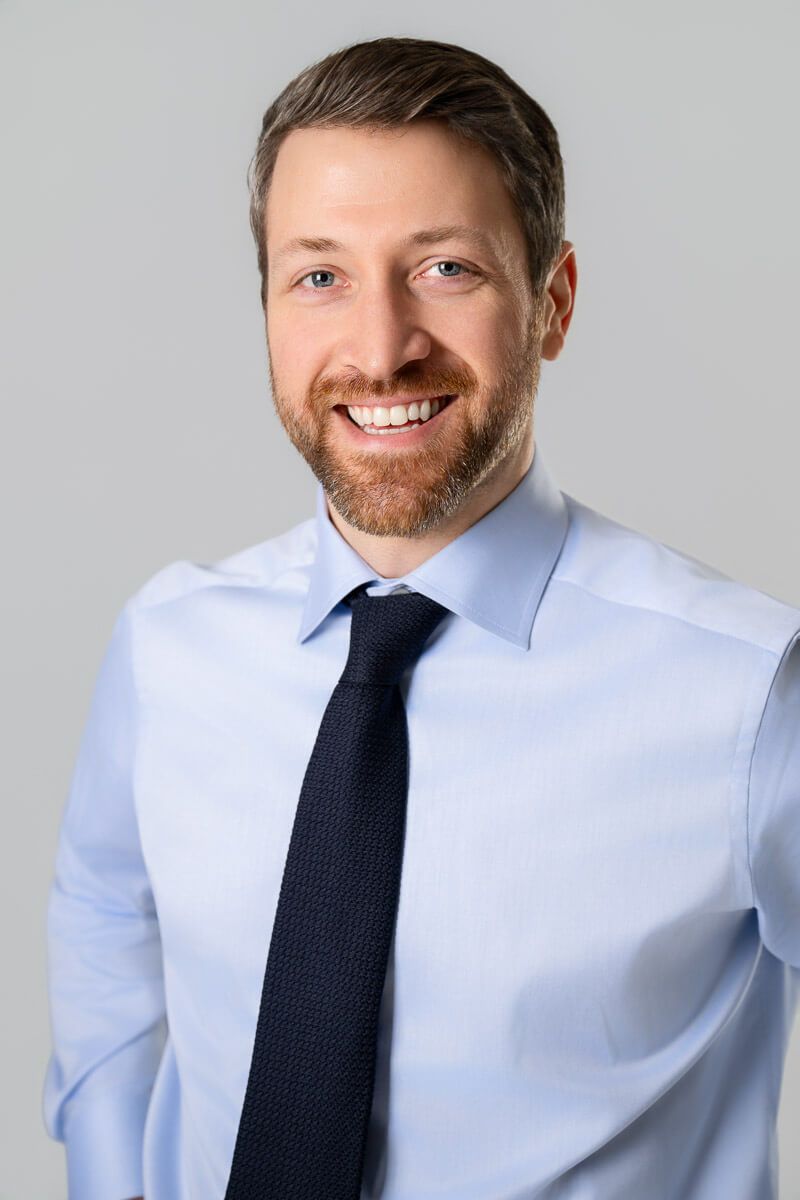 Smiling businessman with beard wearing light blue shirt and dark navy tie against gray background.