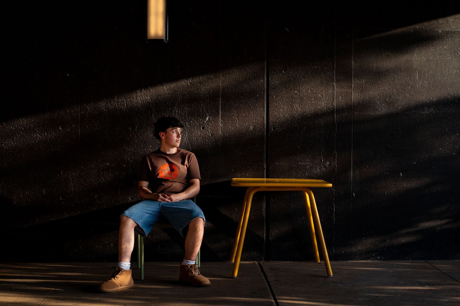 Male senior during a photography session in Downtown Omaha near sunset at Gene Leahy Mall  under the bridge sitting at a yellow table.