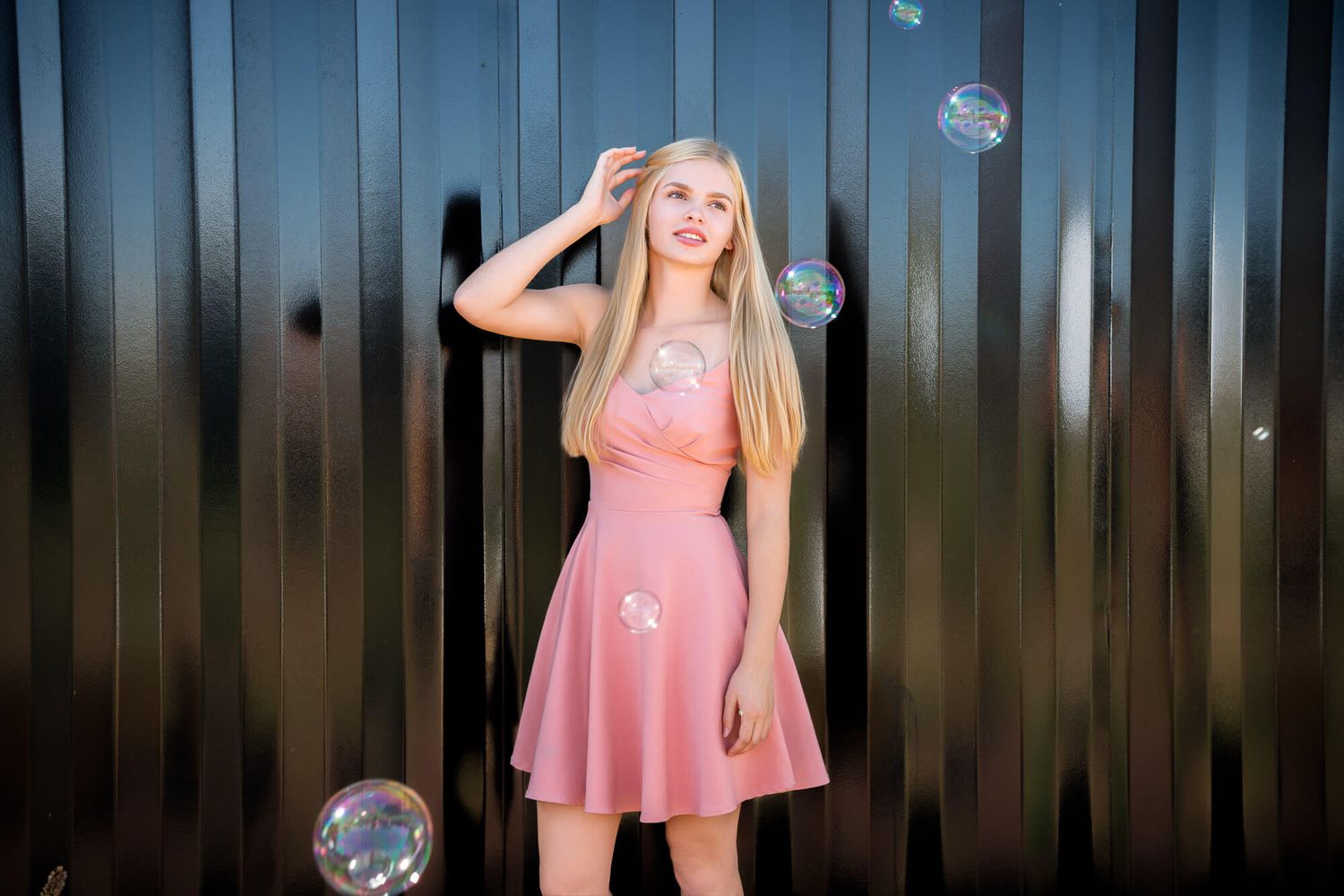 Young blonde woman in pink dress posing gracefully outdoors with colorful soap bubbles floating around her.