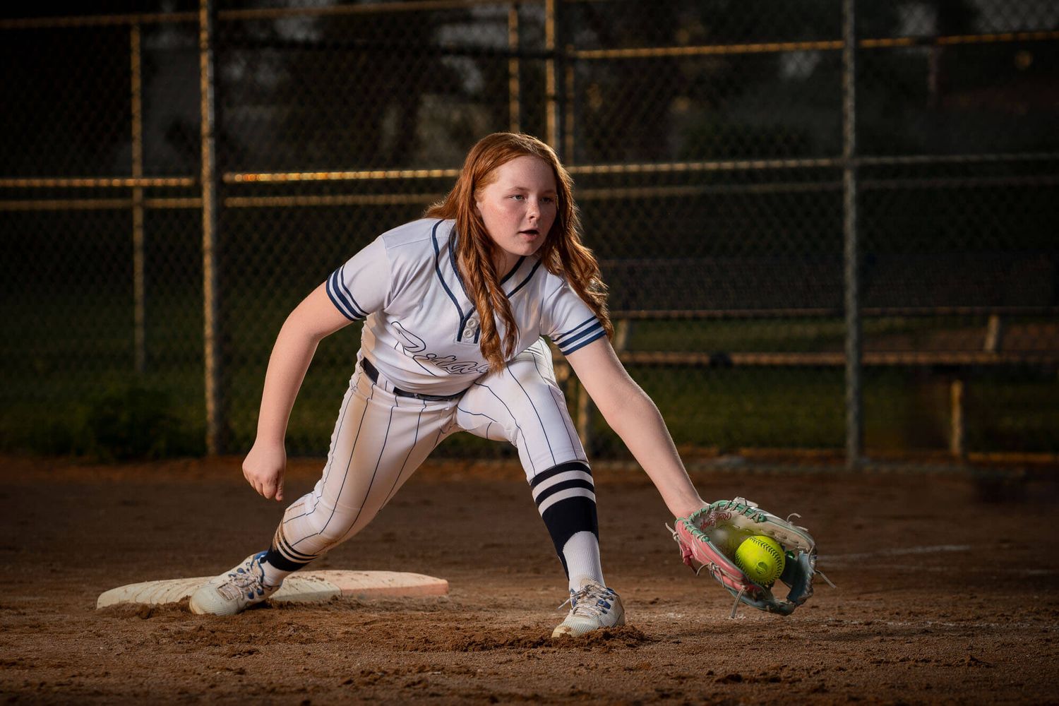 Young female softball player crouching at first base to catch a yellow ball during a night game.
