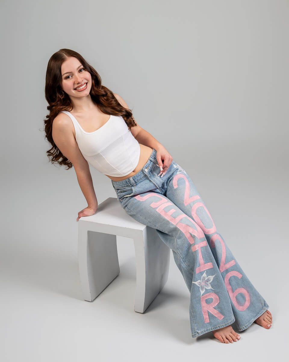 Young woman smiling, wearing white crop top and light blue flare jeans with pink text, seated on white cube.