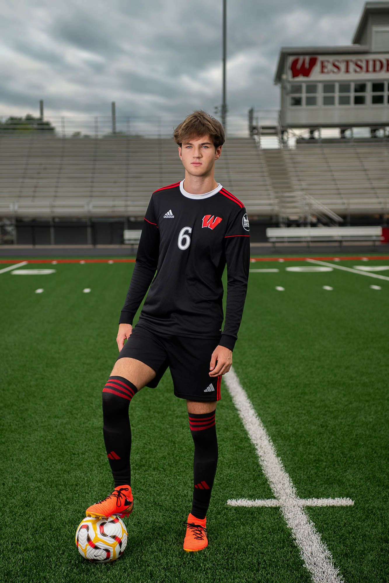 Soccer player kneeling on turf field wearing black uniform with red accents and orange cleats next to soccer ball.