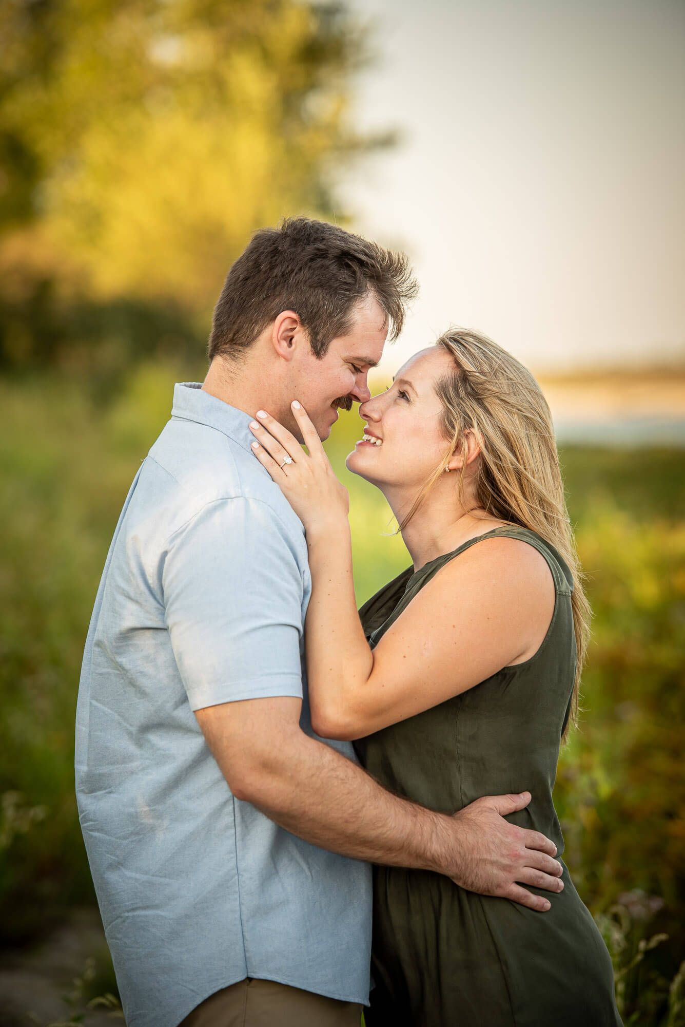 A romantic couple embraces outdoors during golden hour with soft natural lighting and blurred foliage in background.