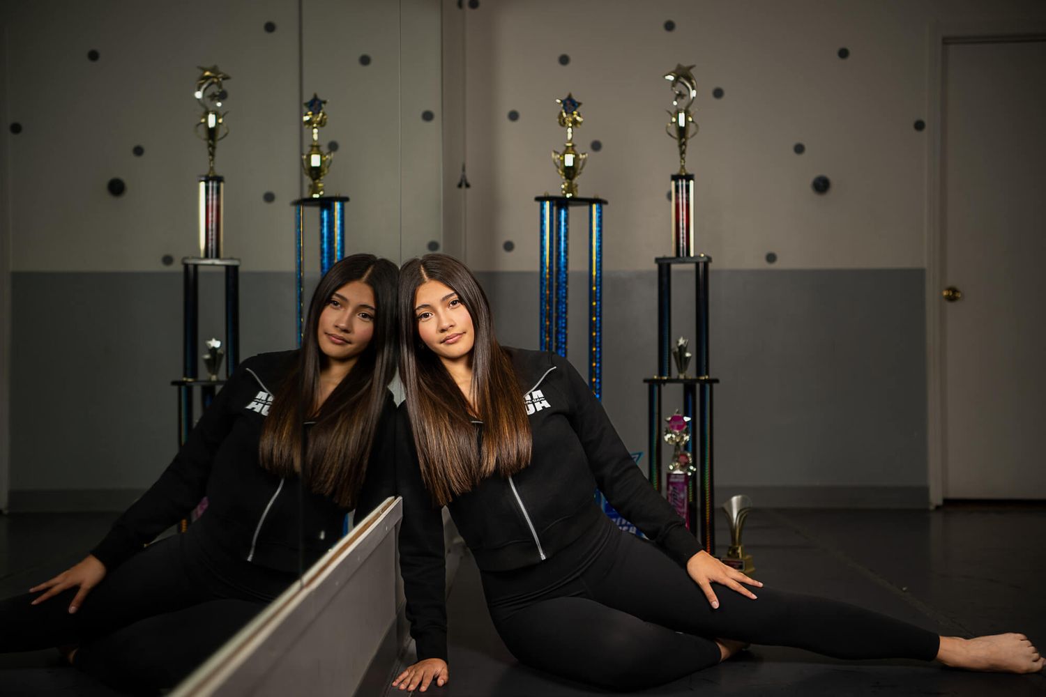Two people in black outfits pose together on a studio floor with lighting equipment visible in the background.