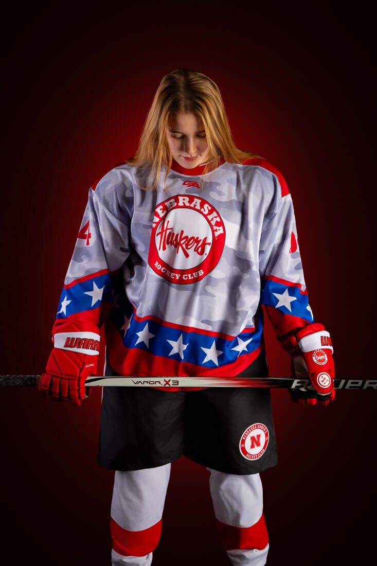 Hockey player poses in white, red and blue uniform with stars and stripes design against dark background.
