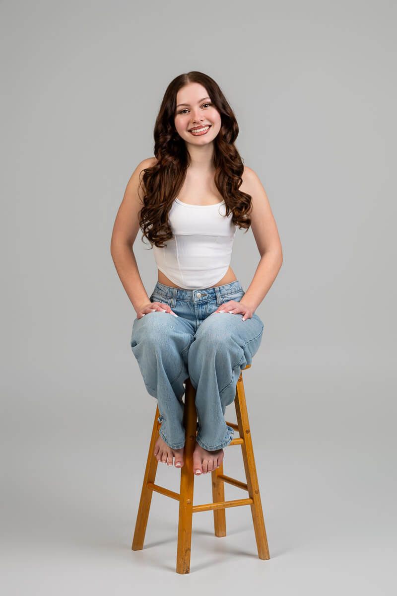 Smiling young woman with long wavy brown hair sitting on wooden stool wearing white crop top and light blue jeans.