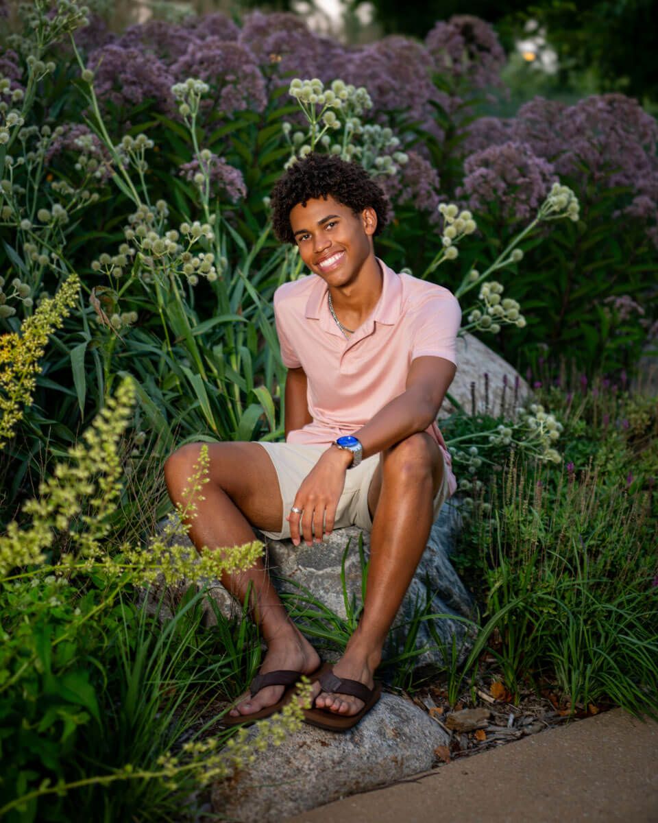 Someone sits casually on a stone wall surrounded by lush garden flowers and greenery at dusk.