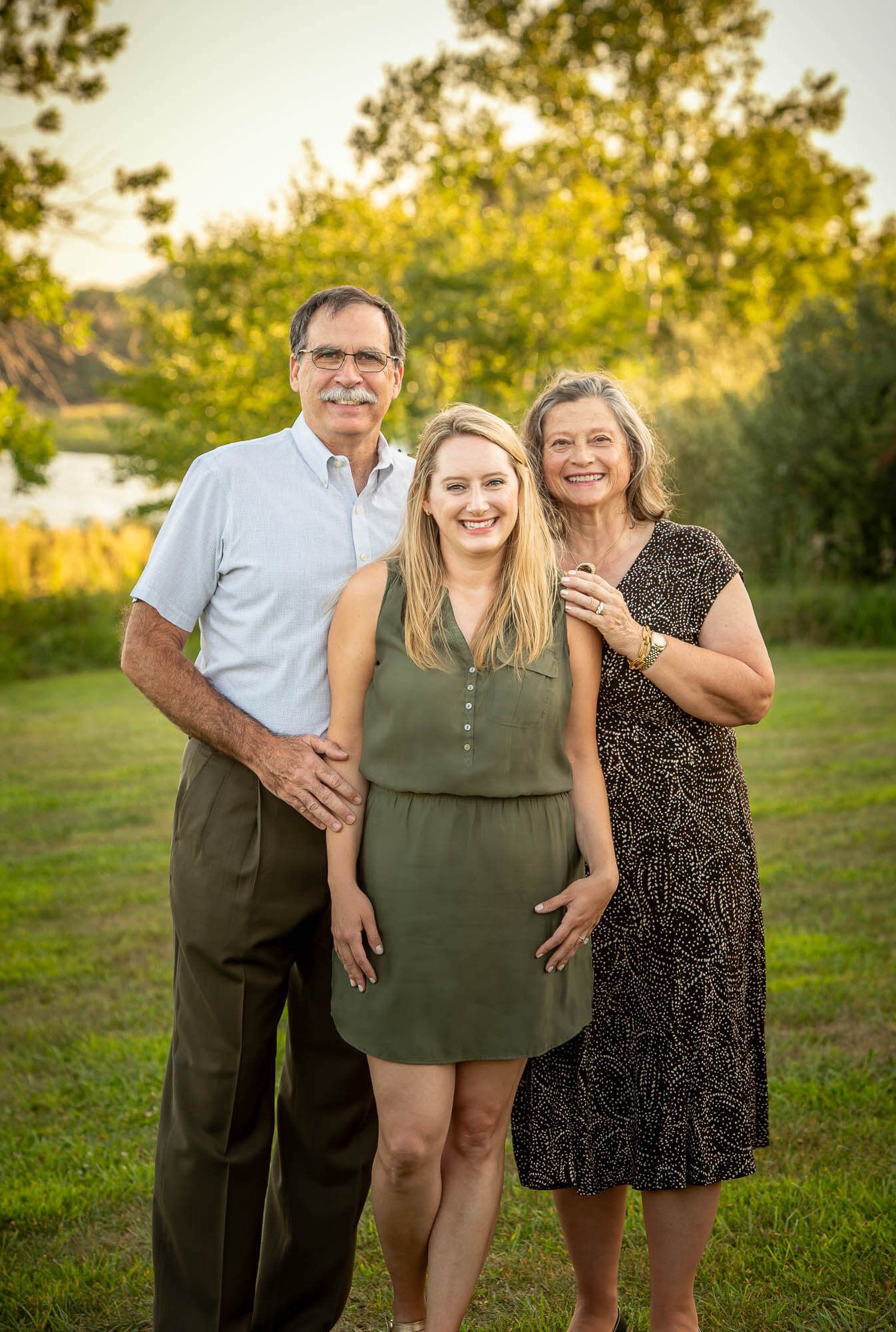 Three people laughing and embracing while standing in a grassy field at sunset.