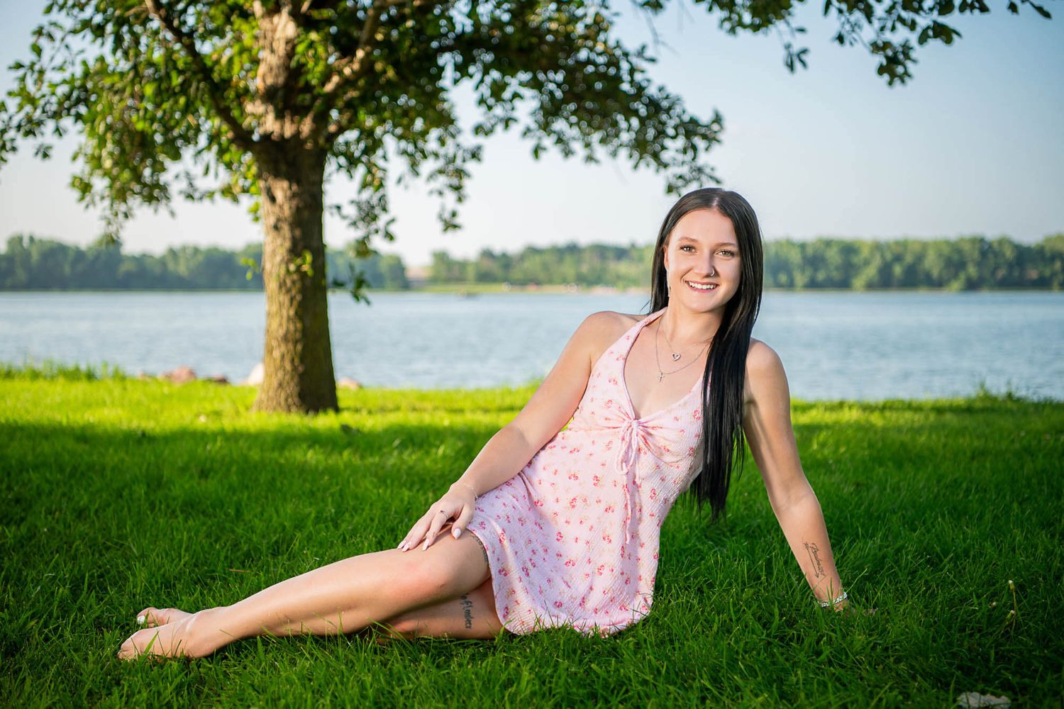 Someone in a pink floral dress relaxes on green grass by a serene lake with a tree in the background.