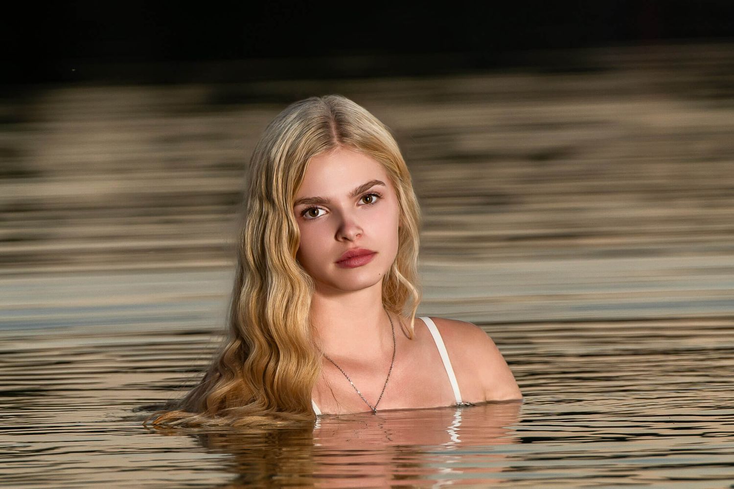 Young blonde woman with long wavy hair emerging from calm water, wearing a white top, in a serene outdoor setting.
