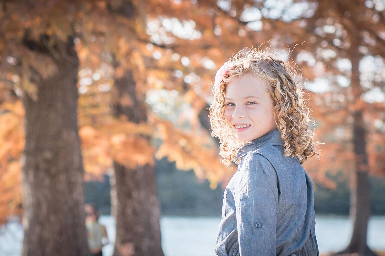 Portrait of curly-haired firl outdoors against autumn trees and lake while taking family photos near Omaha, Nebraska.