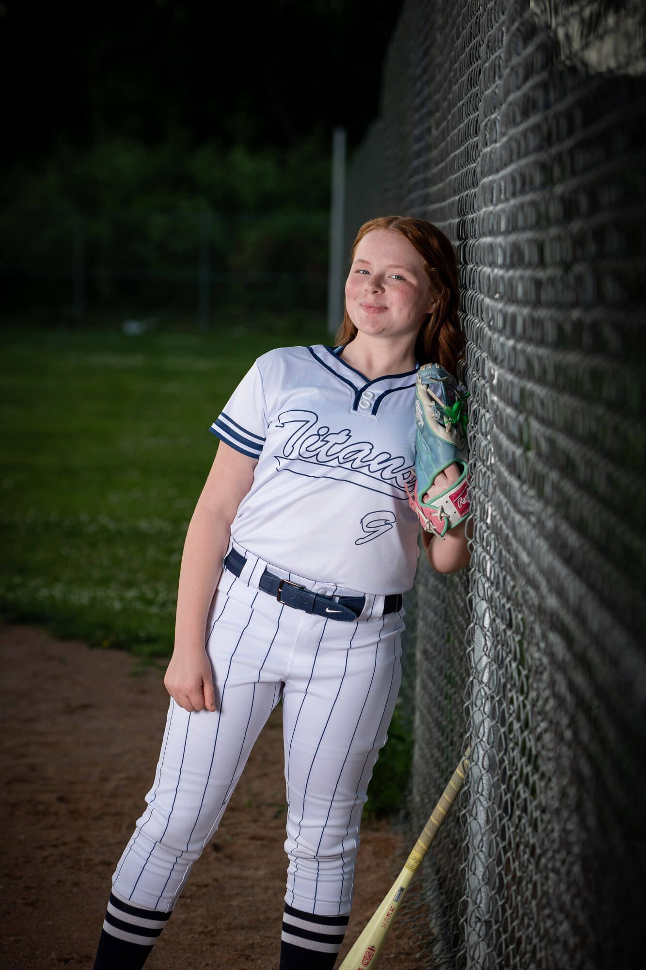 Young girl in Titans softball uniform leaning against brick wall holding glove, portrait photo.