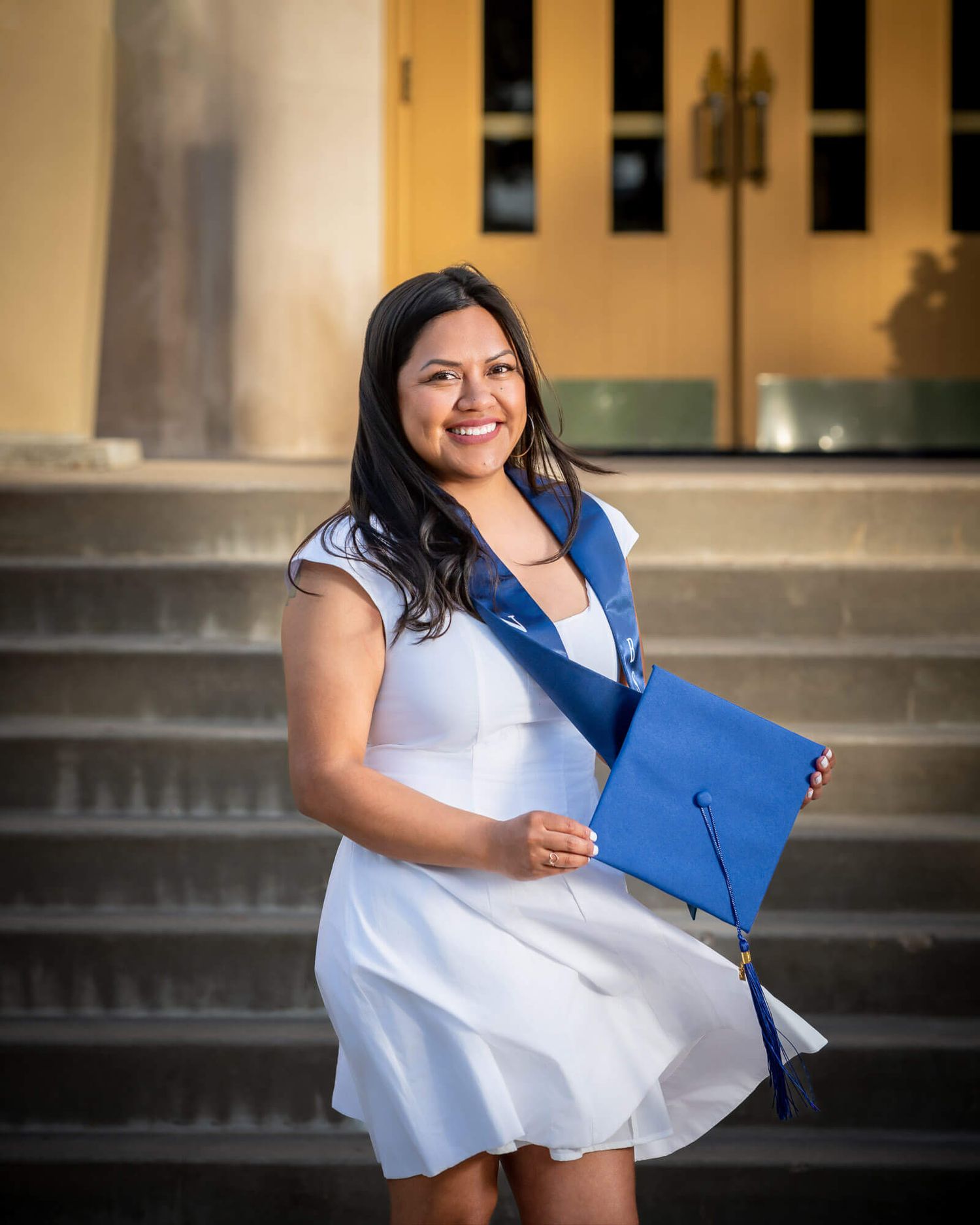 A smiling graduate in a white dress holds a blue cap and diploma on university steps.