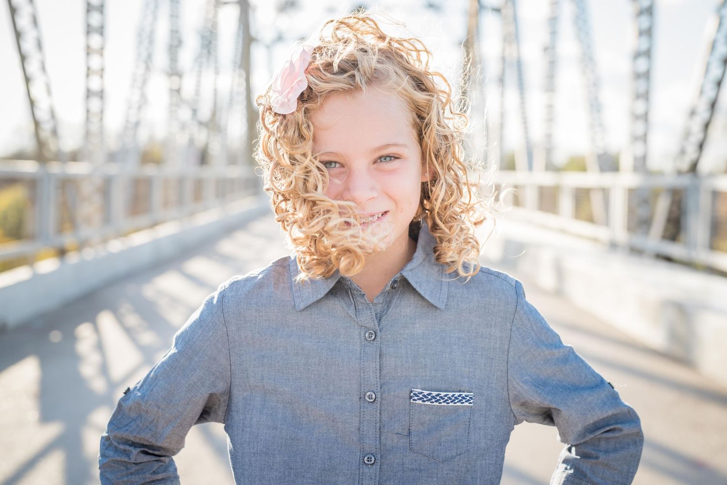 Person in gray button-down shirt stands on metal bridge with curly blonde hair and natural sunlight.