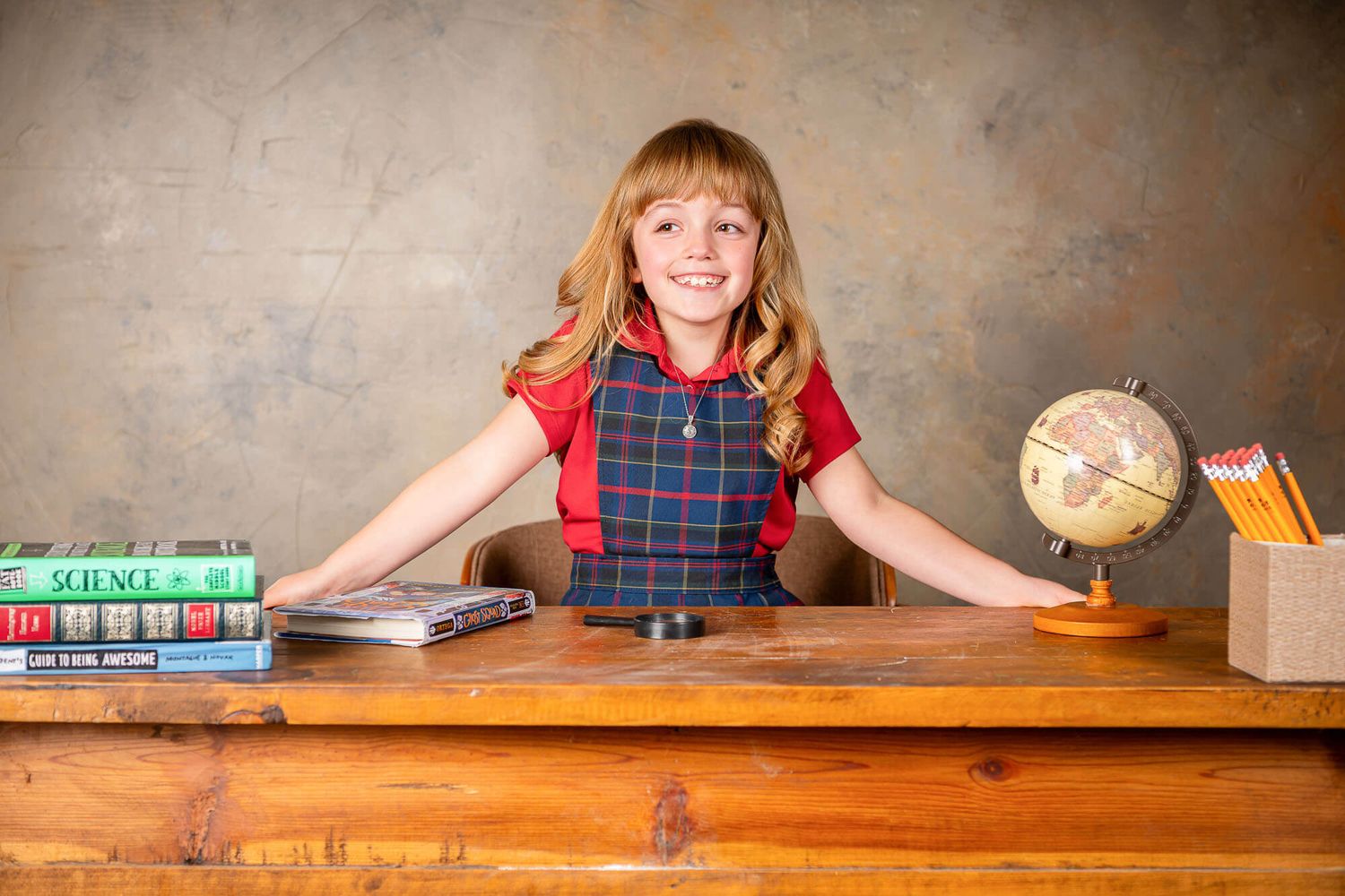 A student in a plaid uniform dress poses with educational materials and a globe on a wooden desk.