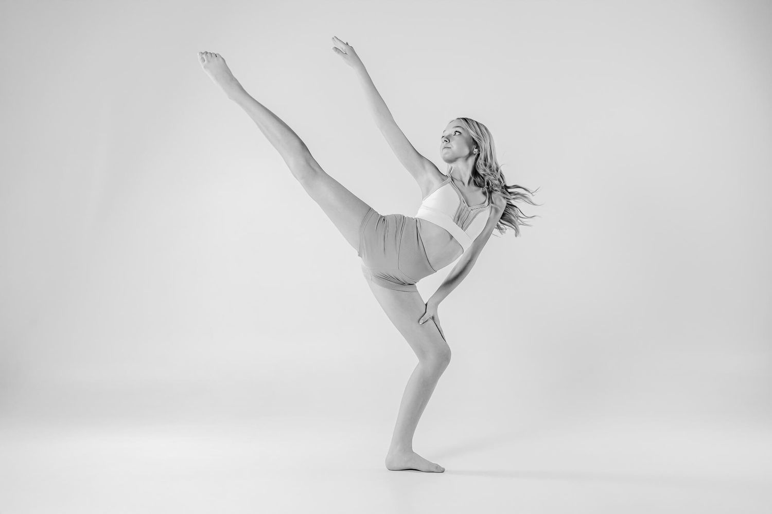 A graceful ballet dancer in white performs an elegant arabesque pose against a light background.