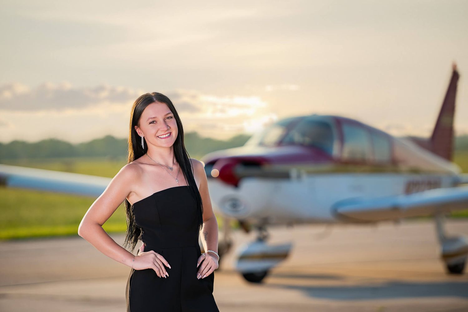 A woman in a black dress poses near small aircraft at sunset on an airfield.