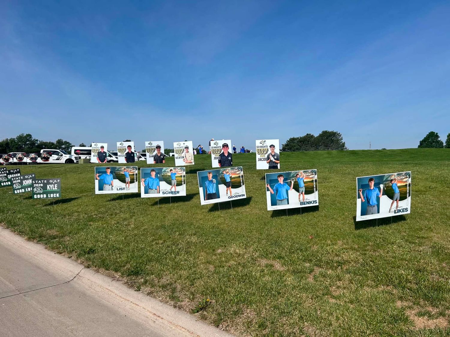 Campaign signs displayed on a green grass field under a clear blue sky at an outdoor political rally event.