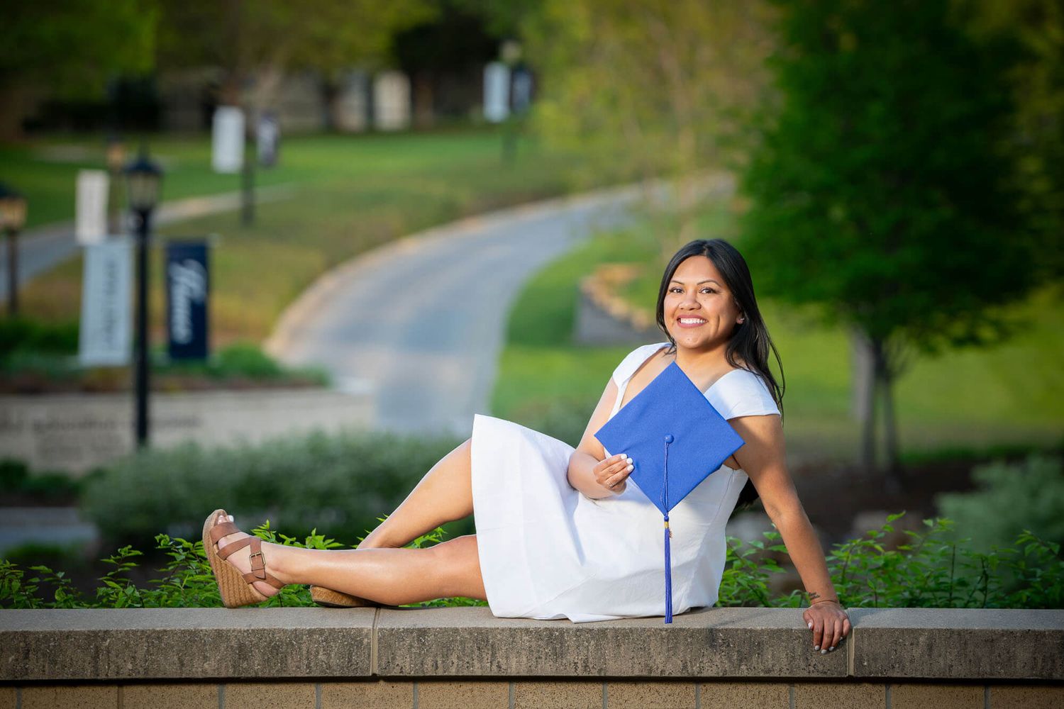 College graduate during photography session taken outdoors on a sunny day with trees and greenery in the background holding cap.