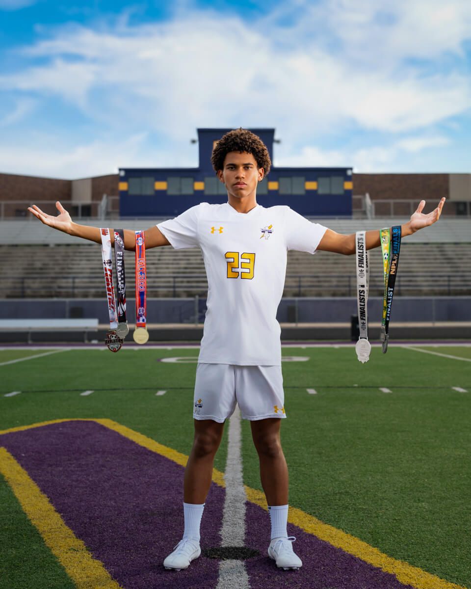 Soccer player #23 in white uniform proudly displays medals on a stadium field, arms outstretched, wearing cleats and socks.