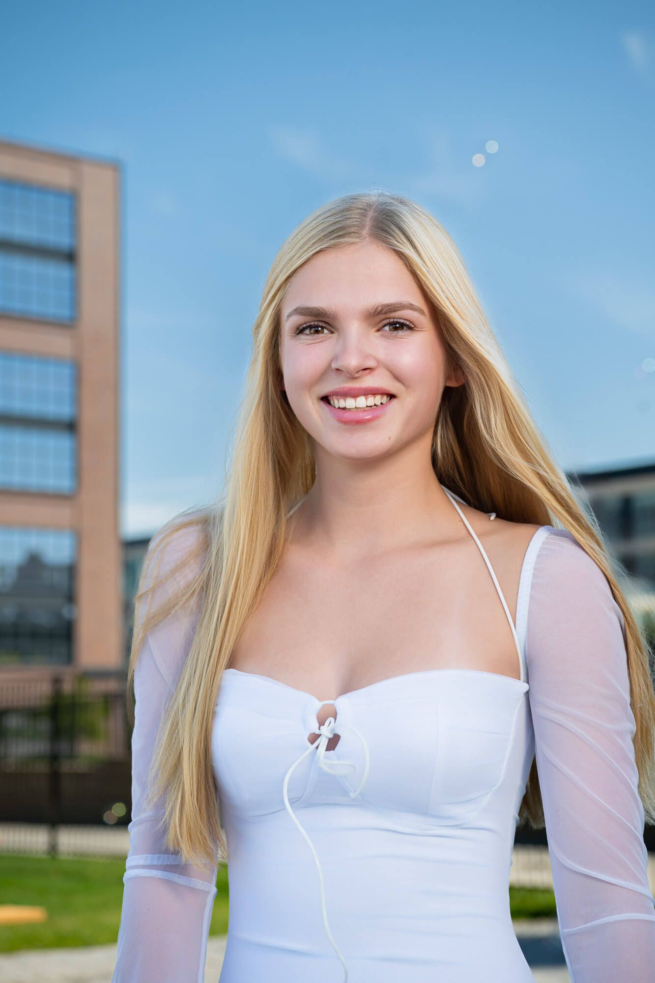 Smiling blonde woman in white dress outdoors with blue sky and building background.