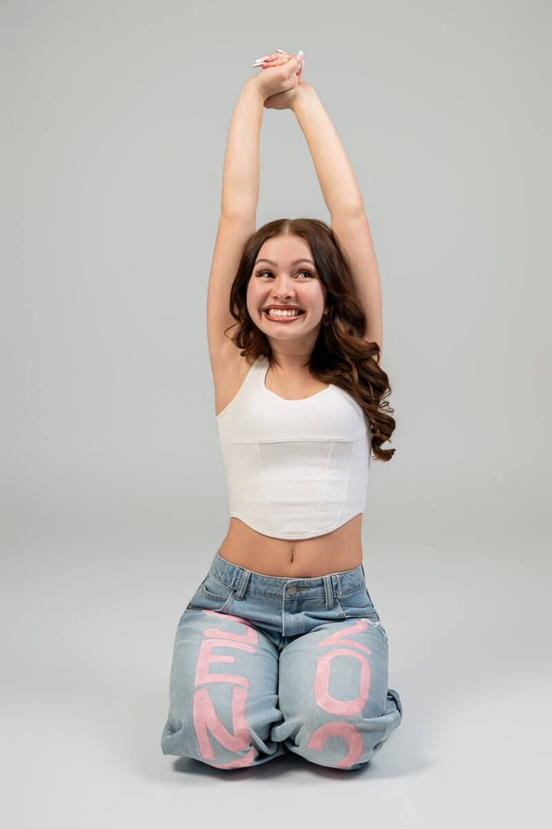 Smiling young woman in white crop top and blue jeans with pink letters, kneeling with arms raised overhead.